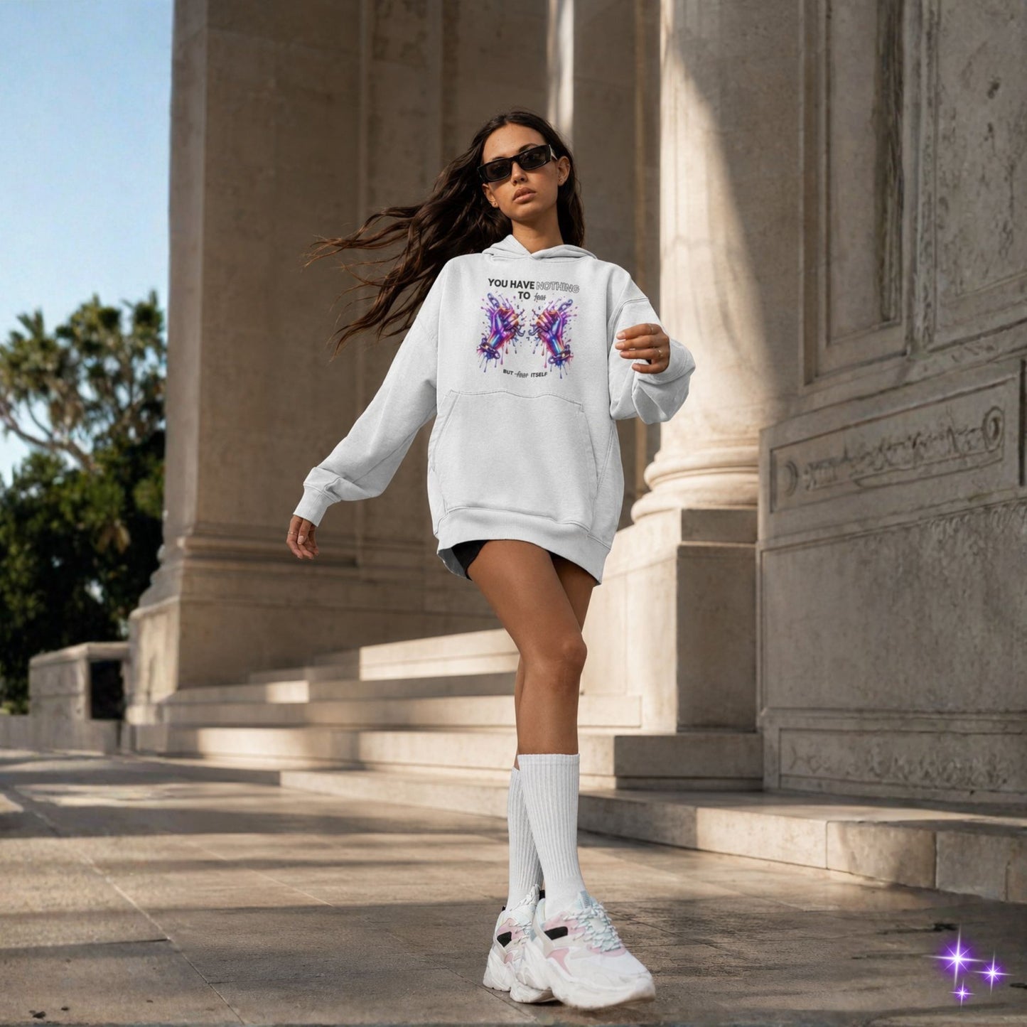 Woman in a white hoodie with butterfly design walking outdoors near classical architecture.