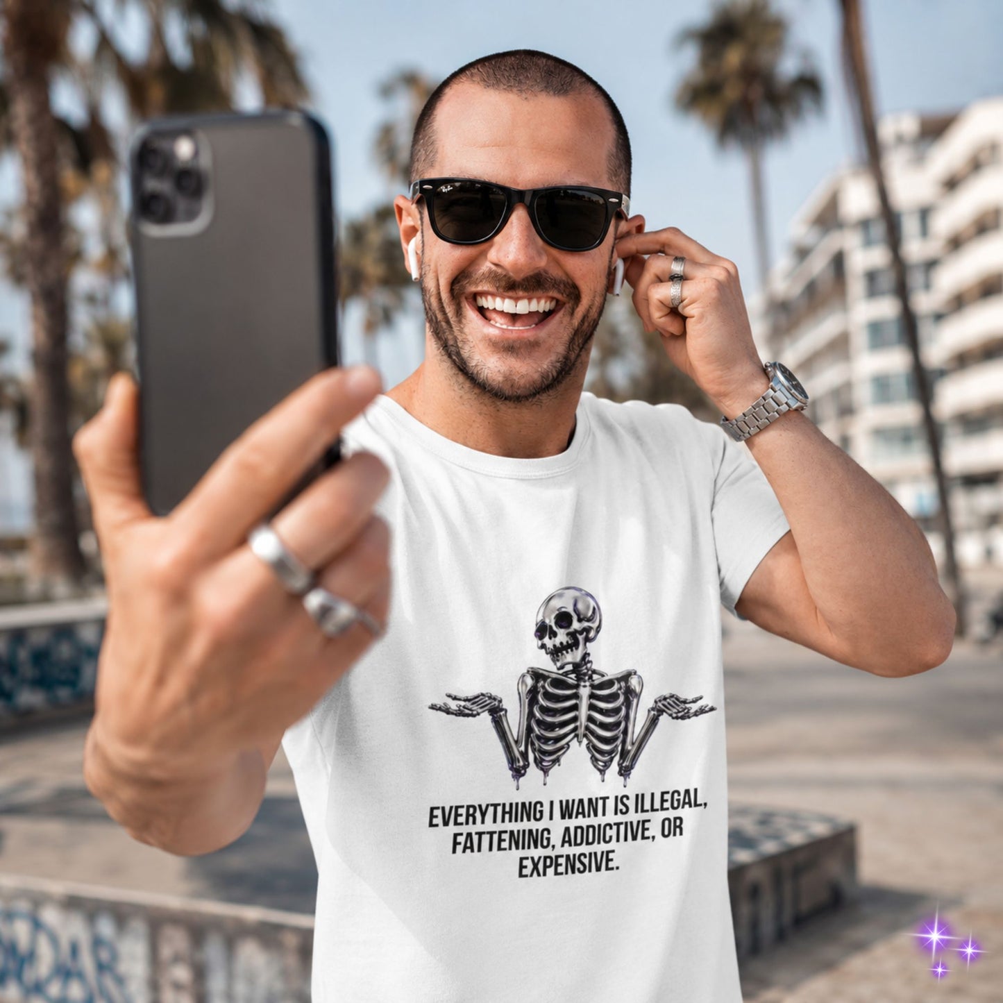 Man taking a selfie wearing a white t-shirt with a skeleton design and text, palm trees and buildings in the background.